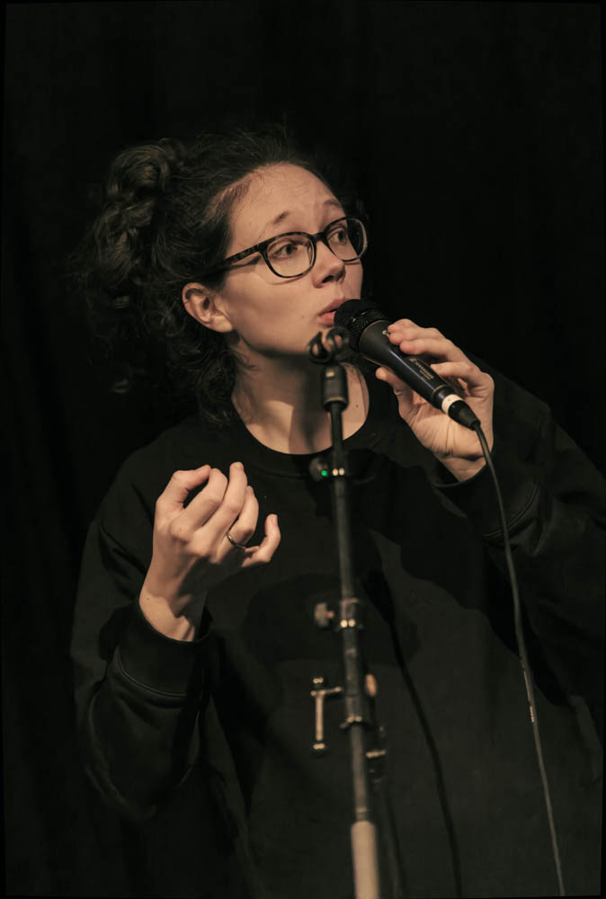 Photo of Tabea singing into a microphone and looking to the side. She is wearing a black sweater against a black background.