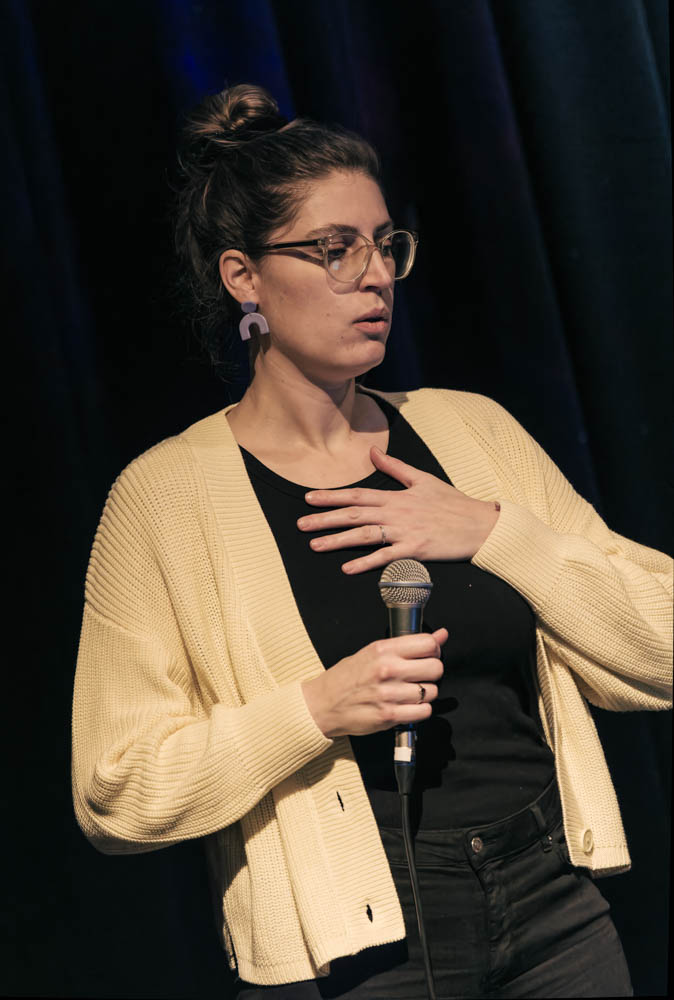 Photo of Ariane holding a microphone and beating on her chest with the other hand. She is wearing black clothing with a beige jacket against a black background.