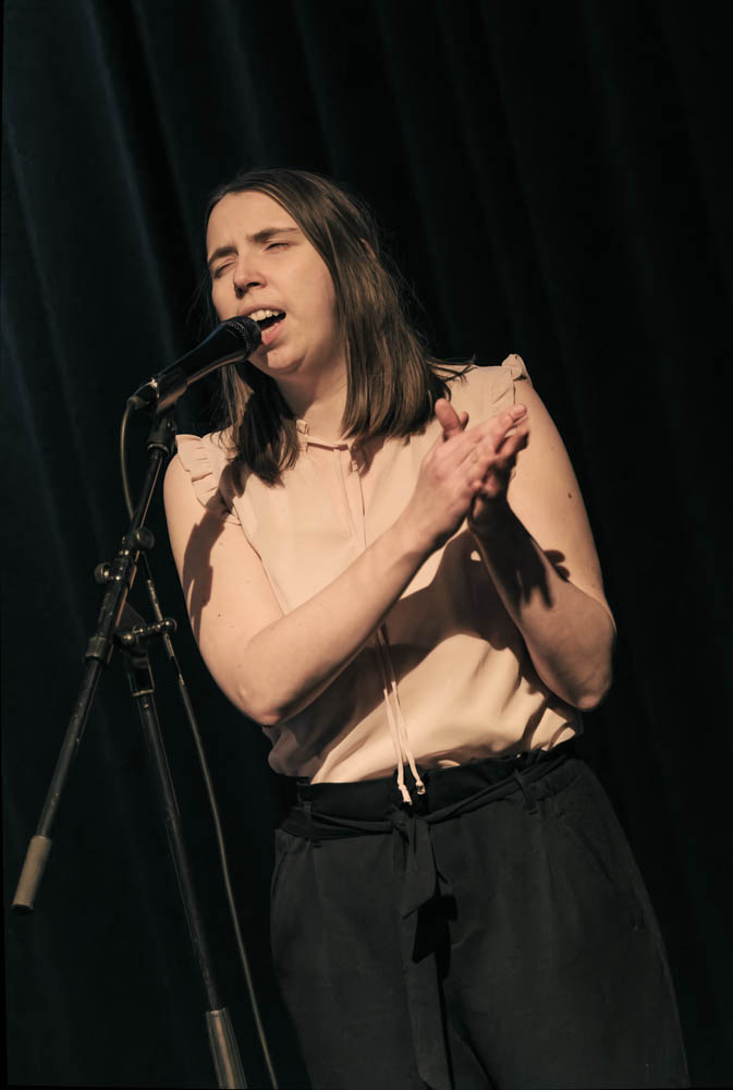 Photo of Lorena singing into a microphone and clapping. She is wearing black pants with a pink blouse against a black background.