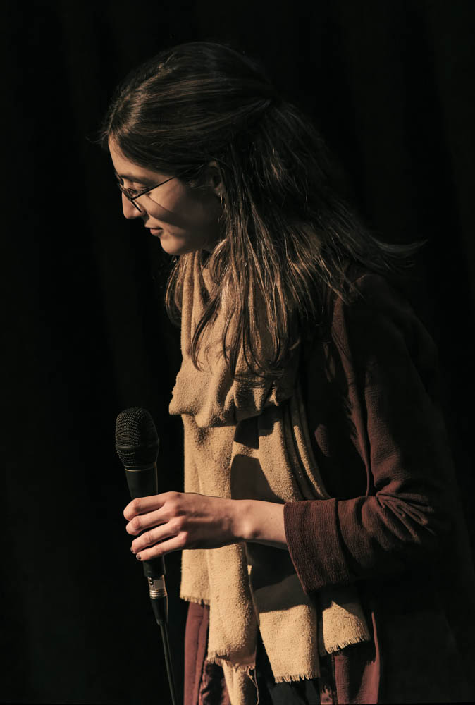 Photo from the side of Marie holding a microphone and looking down to the left with a smile. She is wearing a dark purple jacket with a beige scarf against a black background.