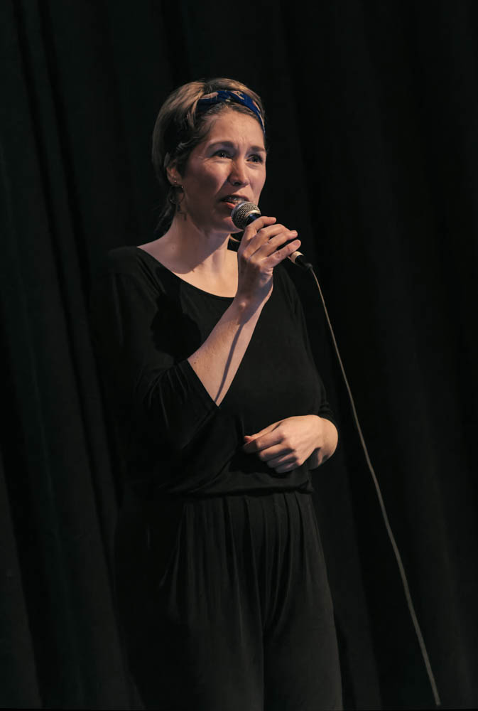 Photo of Mary singing into a microphone in front of a black background. Mary is wearing black clothes.